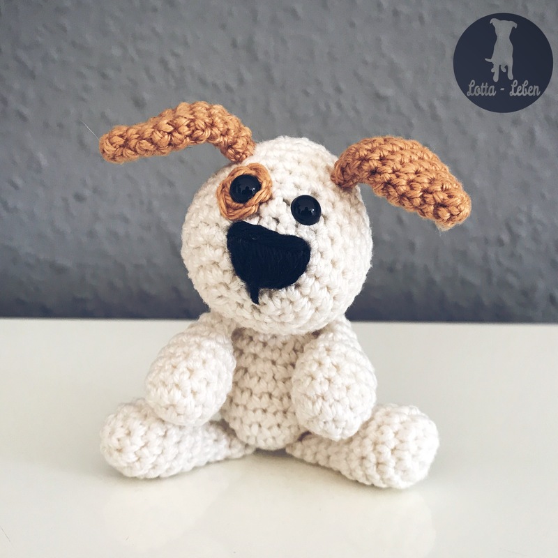 Gehäkelter sitzender Amigurumi-Hund in cremefarbenem Garn mit braunen Ohren, schwarzer Nase und braunem Augenfleck
