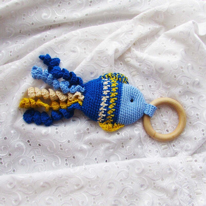 Blue and yellow crocheted fish teether with a natural wooden ring on white eyelet fabric.