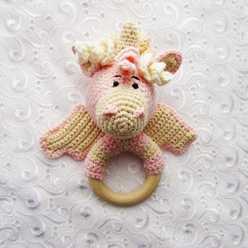 Pink and cream crocheted dragon-shaped baby rattle with a natural wooden ring on a white textured fabric.