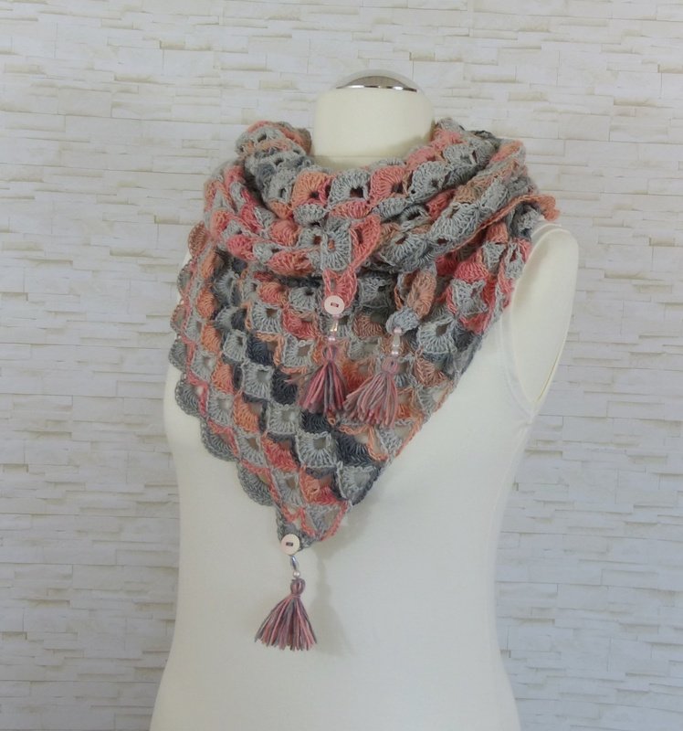 Gehäkeltes dreieckiges Tuch in Grau-, Rosa- und Pfirsichtönen mit Knöpfen und Quasten auf einer Schaufensterpuppe.