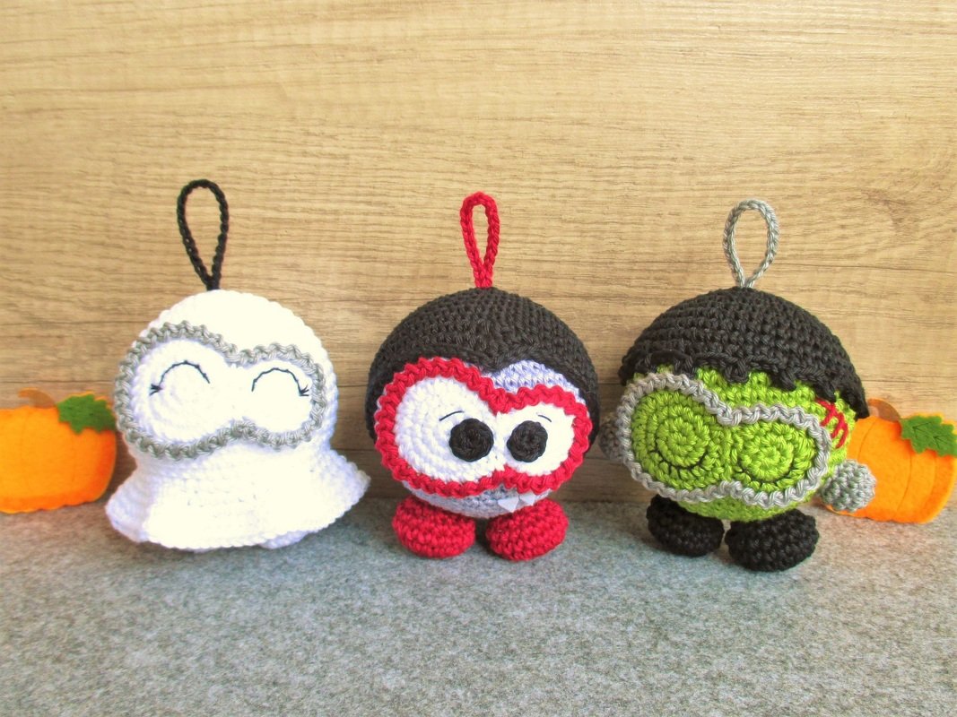 Drei gehäkelte Halloween-Amigurumi: weißer Geist, Vampir mit rotem Rand und grüner Frankenstein mit Aufhängeschlaufen und Kürbissen.