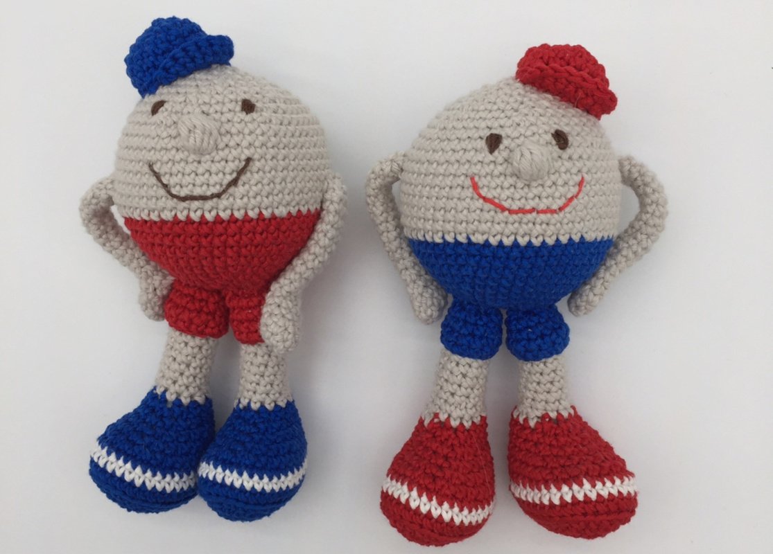 Zwei handgehäkelte Amigurumi-Puppen mit blauer und roter Mütze, kontrastreicher Hose und gestreiften Schuhen auf weißem Hintergrund.