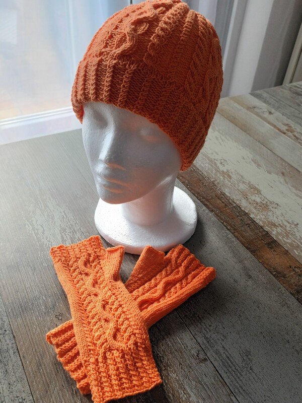 Orangegestrickte Mütze auf Styroporkopf, daneben passende fingerlose Stulpen auf Holzoberfläche.