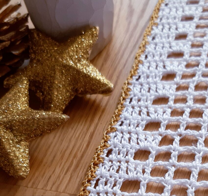 Nahansicht eines weiß gehäkelten Tischläufers mit Gittermuster und goldener Bordüre, daneben goldene Dekosterne.
