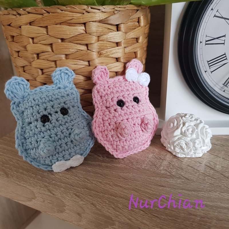 Zwei gehäkelte Nilpferde in Blau und Rosa mit schwarzen Augen und weißen Details, auf einer Holzoberfläche vor einem geflochtenen Korb.