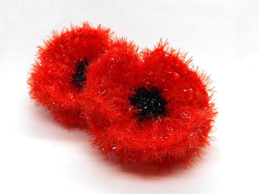 Zwei rote, flauschige gehäkelte Blumen mit schwarzem Zentrum auf weißem Hintergrund