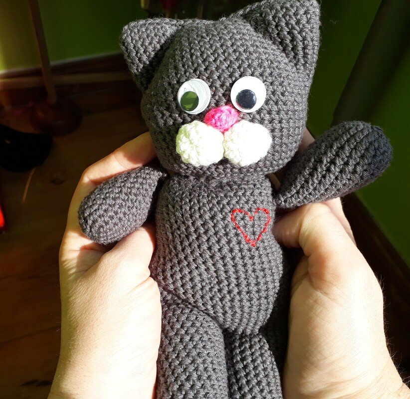 Handgehaltene graue gehäkelte Amigurumi‑Katze mit weißen Schnauzenpolstern, rosa Nase und gesticktem roten Herz