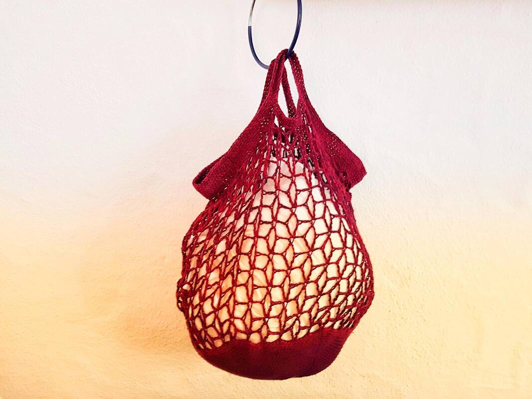 Bordeauxrote gehäkelte Netztasche mit offenem Maschenmuster, hängend an einem Haken vor einer hellen Wand