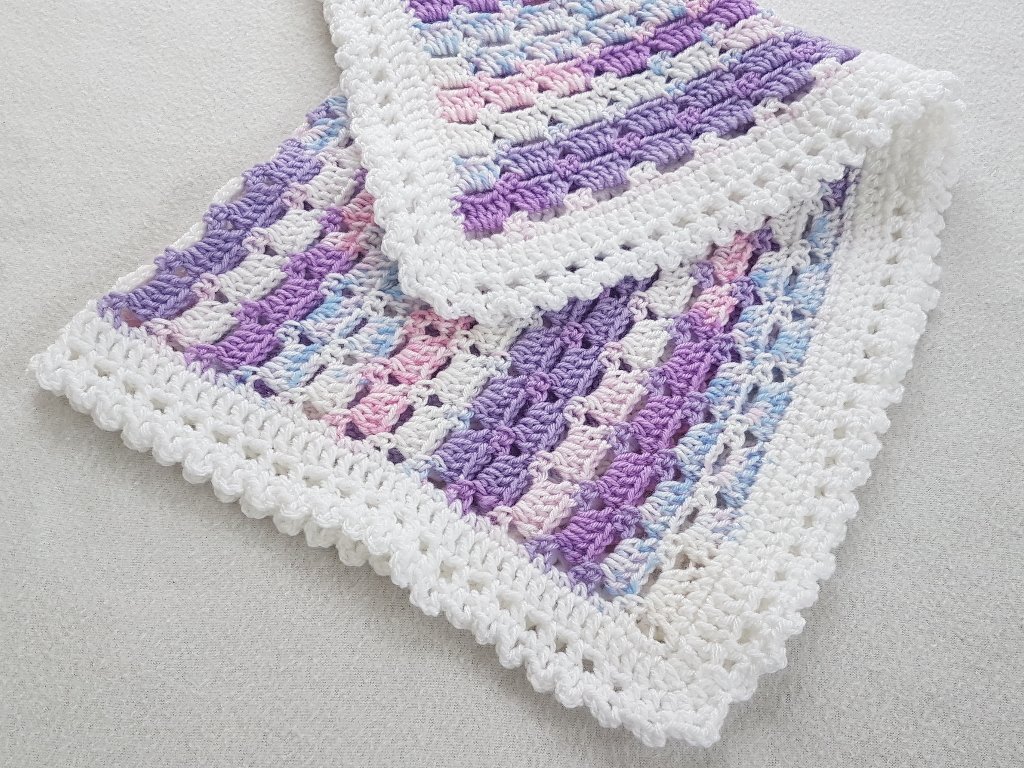 Gehäkelte Babydecke mit lila, rosa und hellblauen Streifen sowie weißer Muschelbordüre