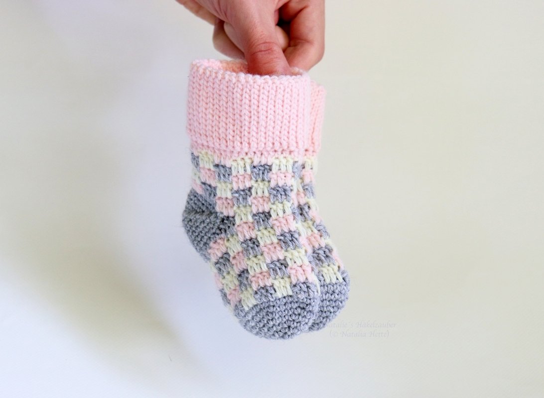 Hand holding a single crocheted baby sock with a pink ribbed cuff and a pink, cream, grey checkerboard pattern.