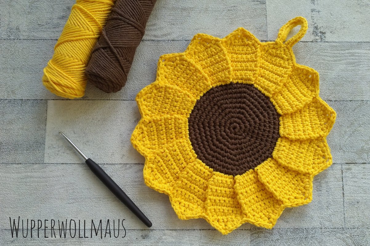 Fröhlicher gehäkelter Topflappen in Sonnenblumenform mit gelben Blättern und braunem Zentrum, daneben Garn und Häkelnadel
