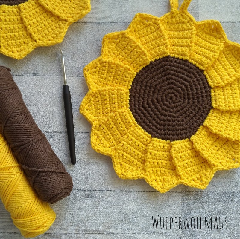 Gehäkelter Sonnenblumen-Topflappen mit gelben Blütenblättern und braunem Zentrum, mit Garn und Häkelnadel auf Holz.