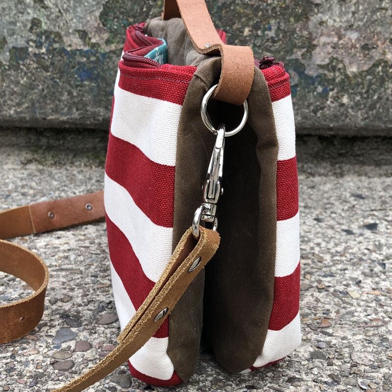 Rot-weiß gestreifte Tasche mit braunen Seiten, abnehmbarem Ledergurt und Metallkarabiner auf Beton.