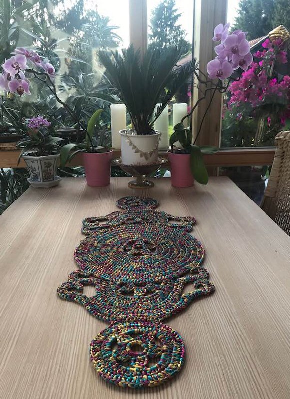 Langer gehäkelter Tischläufer aus buntem Garn mit detailreichem Muster auf hellem Holztisch, umgeben von Pflanzen