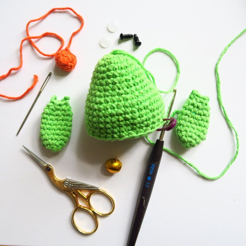 Materialien für Amigurumi: grüne gehäkelte Teile, orange Wolle, Häkelnadel, Schere, Sicherheitsaugen und kleine Glocke.