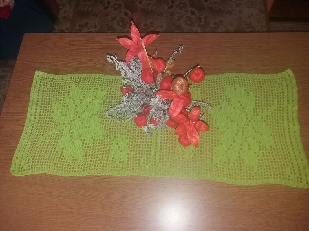 Hellgrüner gehäkelter Tischläufer im Filethäkelmuster mit Blattmotiven und herbstlicher Dekoration aus Lampionblumen auf einem Holztisch.