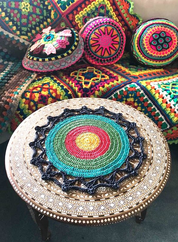 Boho-Häkelarrangement: rundes, buntes Deckchen auf Tisch vor gehäkelter Decke und bunten Kissen