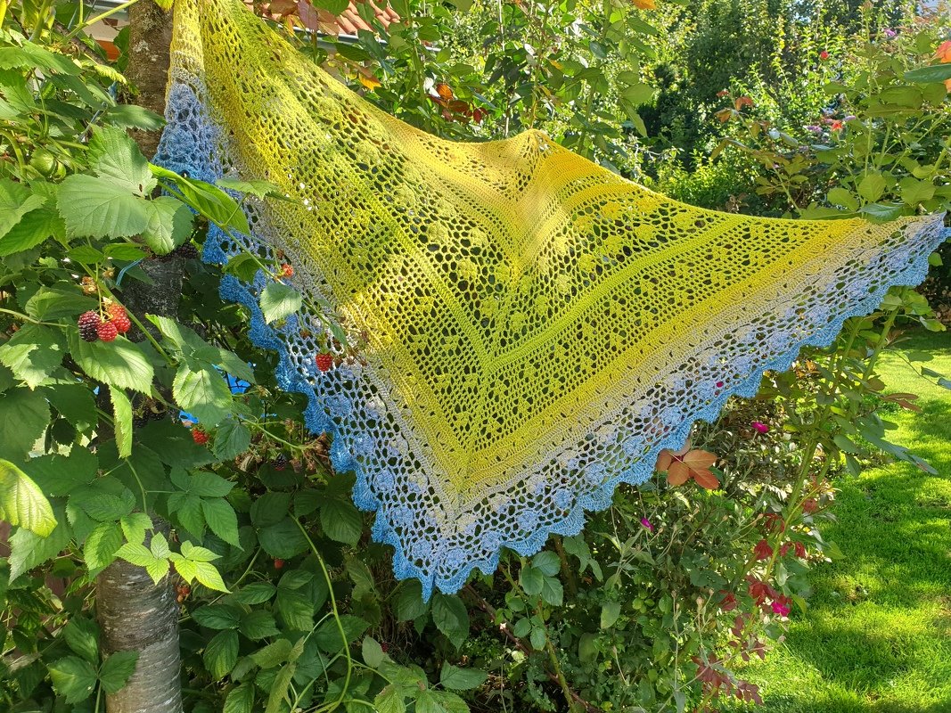 Gehäkeltes Dreieckstuch mit Gelb‑Blau‑Farbverlauf und Lochspitze, an einem Ast im Garten aufgehängt.