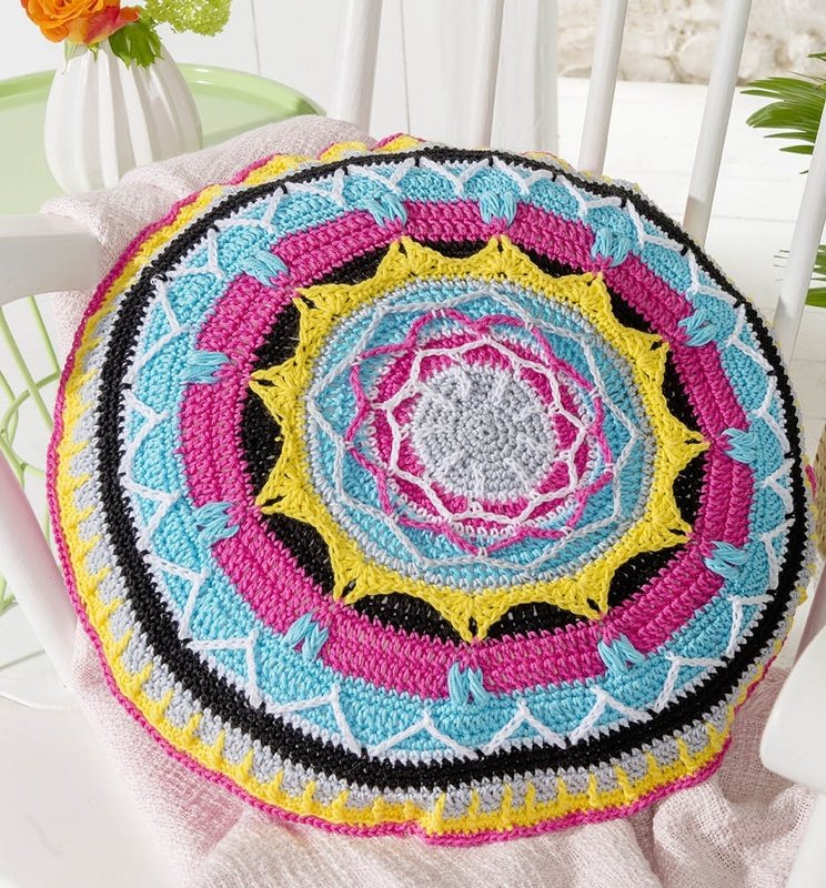 Rundes, gehäkeltes Kissen mit komplexem Mandala-Muster in Pink, Türkis, Gelb und Schwarz auf einem weißen Stuhl.