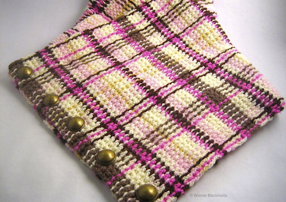 Nahaufnahme eines gehäkelten Karomusters in Rosa, Braun und Creme mit fünf messingfarbenen Knöpfen.