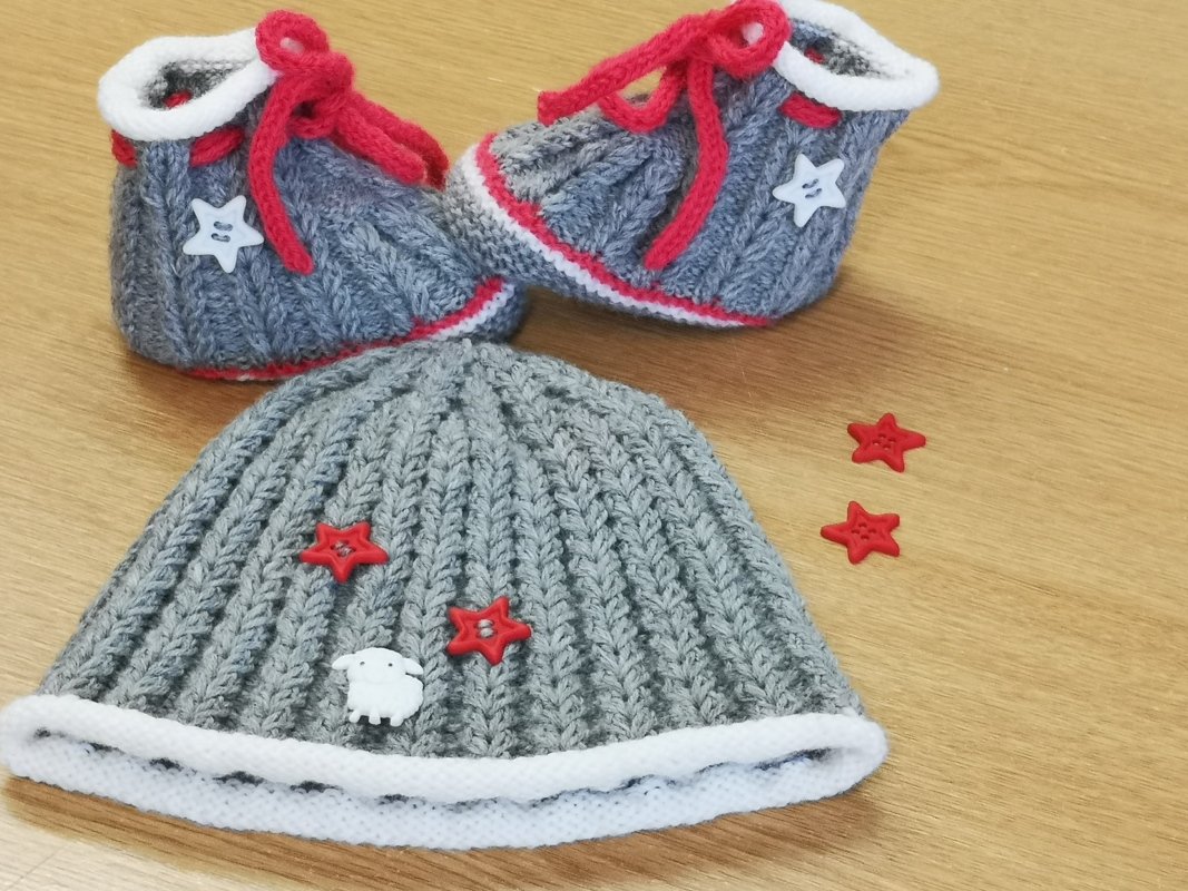Grau gestricktes Babymützen-Set mit Booties, Stern- und Schafknöpfen sowie roten Schnürsenkeln auf hellem Holztisch.