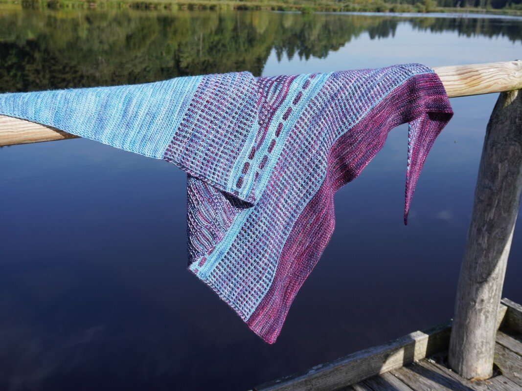 Gestricktes Dreieckstuch in Blau- und Violetttönen mit Loch- und Rippenmuster, über einem Holzgeländer am See drapiert.