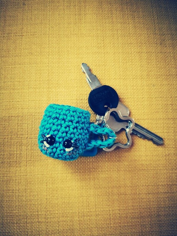 Türkis gehäkelte Mini-Tasse mit schwarzen Augen an Schlüsselring auf gelbem Hintergrund