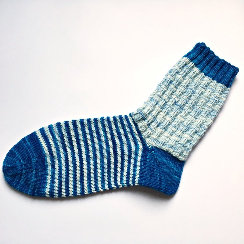Gestrickte Socke in Blau und Weiß mit quer gestreiftem Fuß und geripptem Schaft auf weißem Hintergrund.