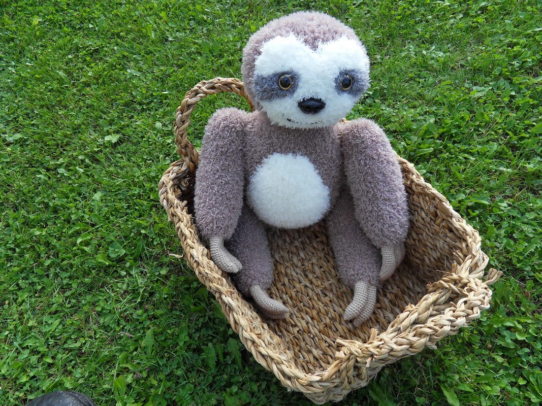 Amigurumi-Faultier mit braun-weißem Fell in einem Weidenkorb auf grüner Wiese