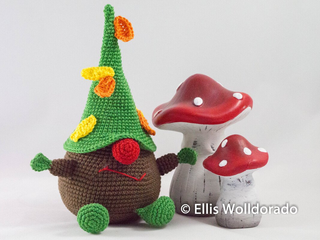 Braunes, rundes Amigurumi mit grünem Spitzhut und zwei rot-weiß gepunkteten Pilzen