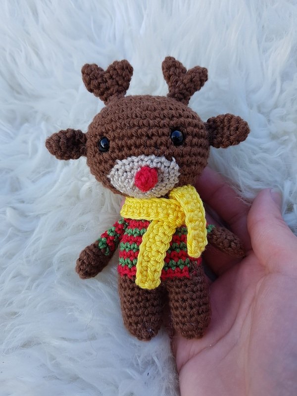 Braunes gehäkeltes Amigurumi-Rentier mit roter Nase, rot-grün gestreiftem Pullover und gelbem Schal, in einer Hand gehalten