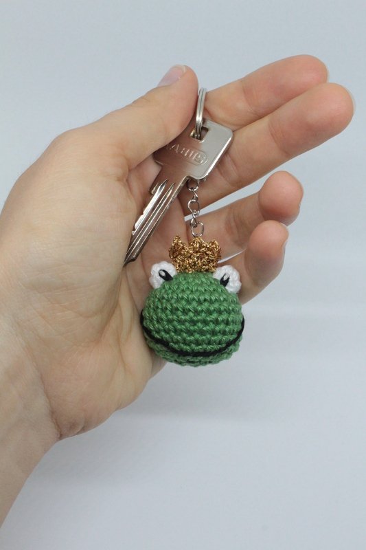 Hand hält einen Schlüssel mit gehäkeltem grünen Frosch‑Schlüsselanhänger mit goldfarbenem Krönchen.