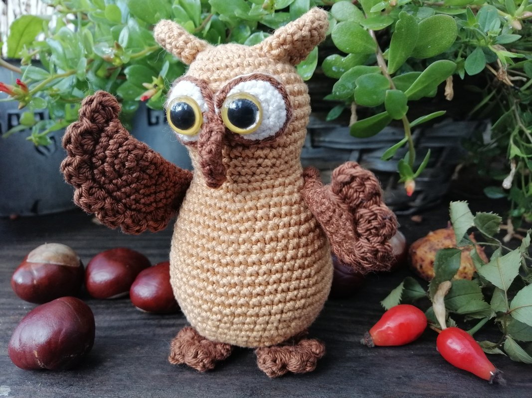 Gehäkelte Amigurumi-Eule beige mit braunen Flügeln und großen Augen auf Holzbrett zwischen Kastanien und Hagebutten