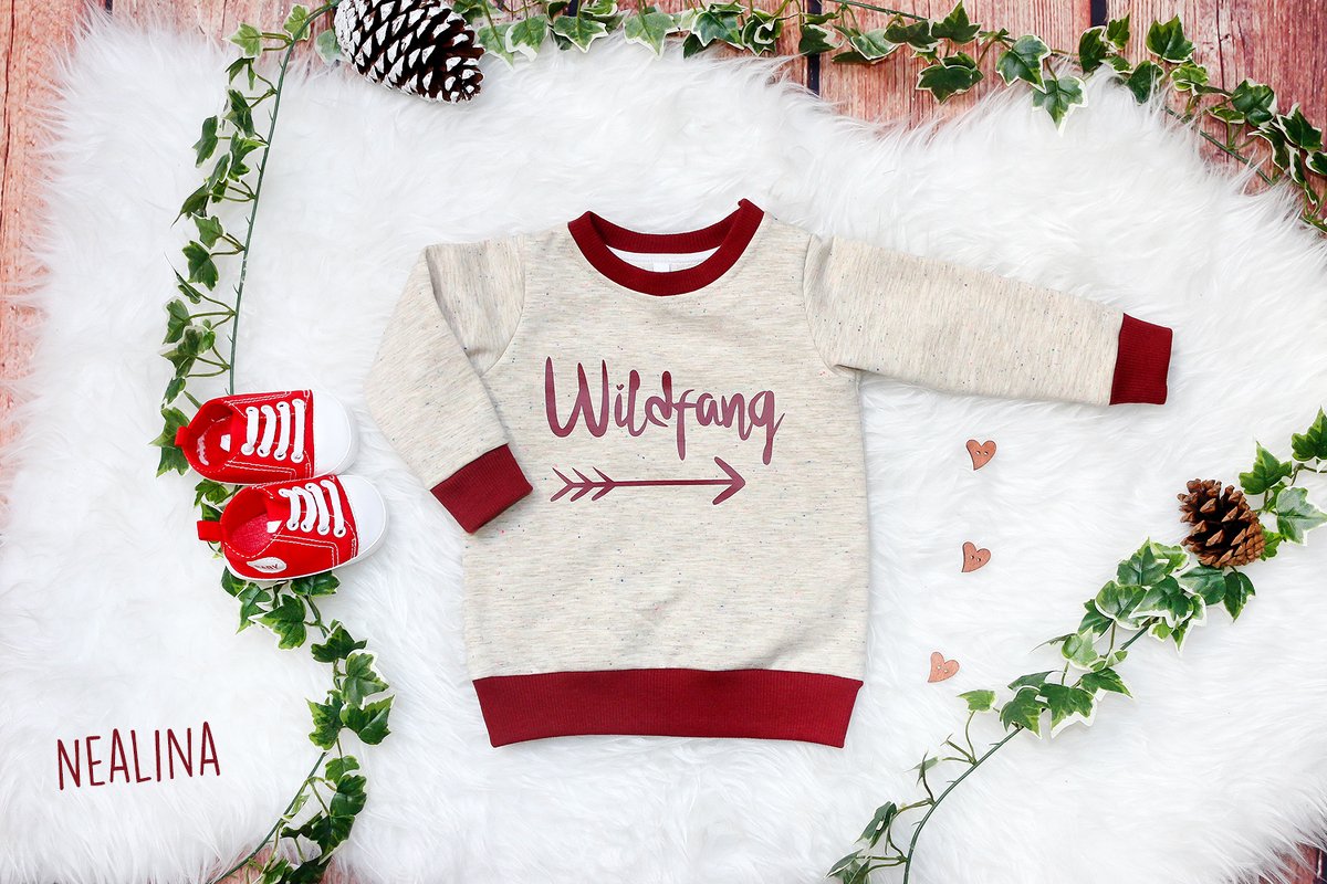 Beiges Baby‑Sweatshirt mit dunkelrotem Bündchen und Plottmotiv ‚Wildfang‘ mit Pfeil, arrangiert mit roten Babyschuhen und Efeuranken auf hellem Felluntergrund.