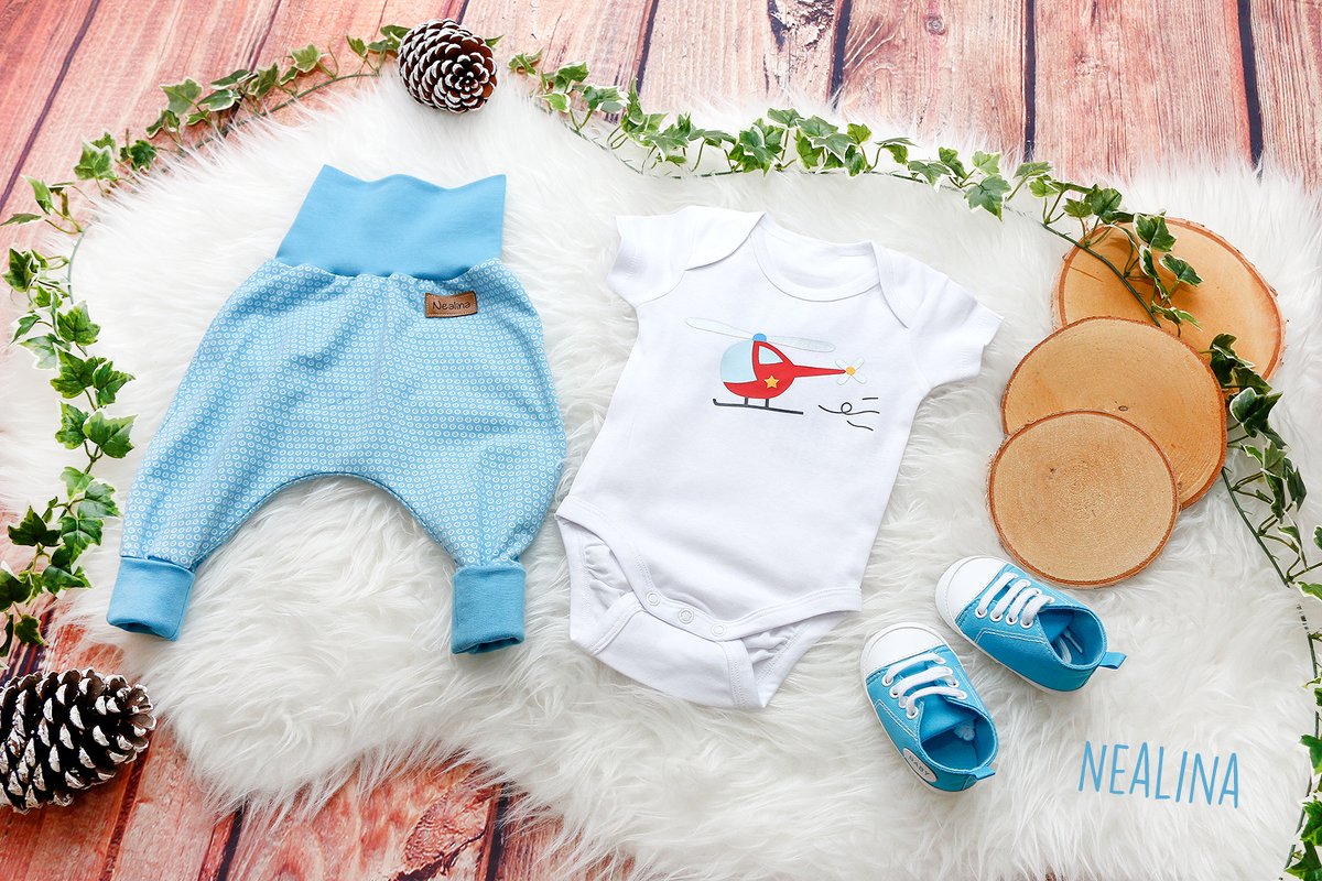 Baby-Body mit Hubschrauber-Applikation, gemusterte Pumphose und Babyschuhe auf flauschigem Untergrund.
