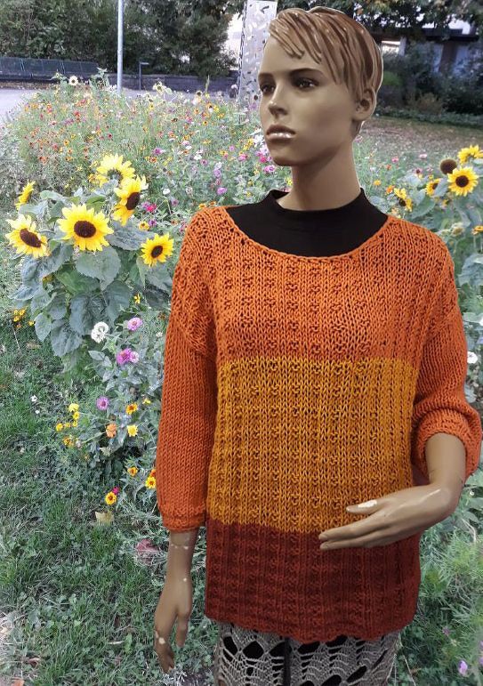 Mannequin trägt einen gestrickten Pullover in Orange, Gelb und Rostrot mit Rundhalsausschnitt und 3/4-Ärmeln vor einer Blumenwiese.