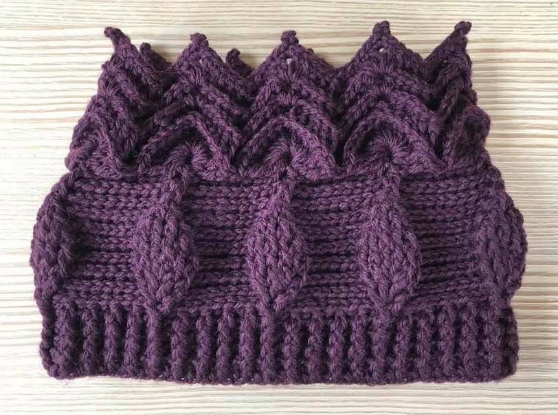 Dunkellila gehäkelte Mütze mit Zopfmuster, Rippenbündchen und gezacktem Rand auf hellem Holzuntergrund