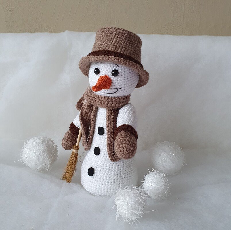Handgehäkelter Amigurumi-Schneemann mit braunem Zylinder, Schal und Besen, umgeben von weißen Dekoschneebällen.
