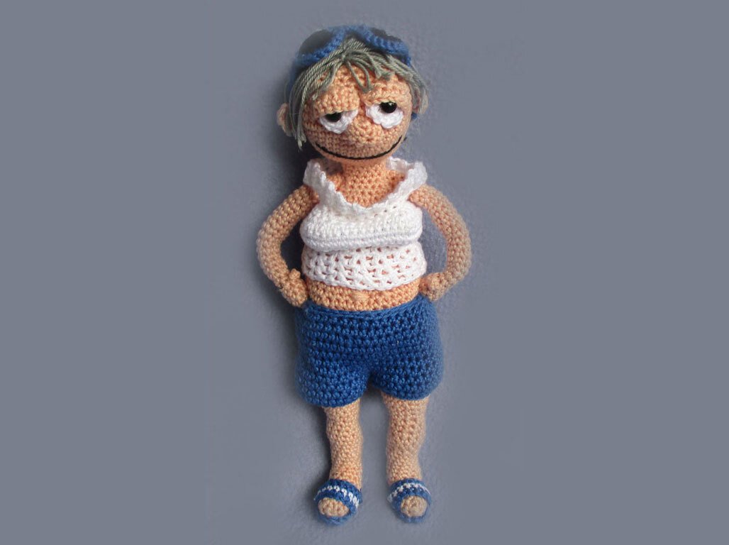 Crocheted amigurumi doll in white ruffled top and blue shorts with sandals on grey background
