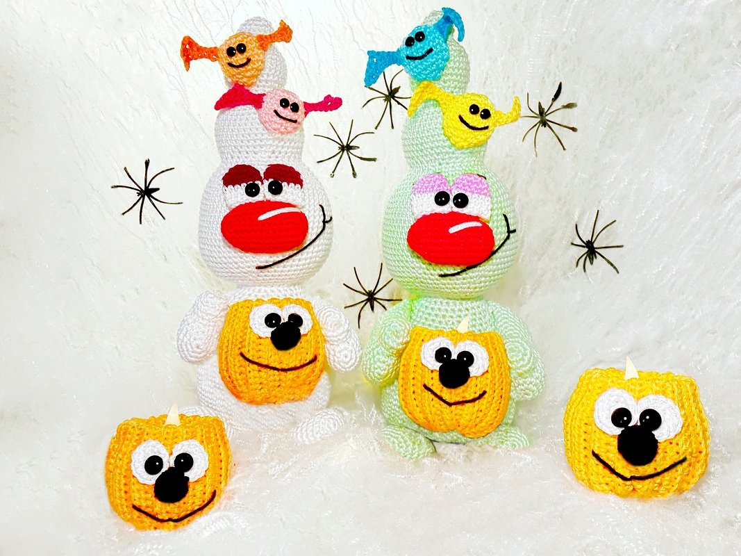 Zwei gestapelte gehäkelte Amigurumi-Figuren (weiß und hellgrün) mit roten Nasen, vorn je ein gehäkelter Kürbis und kleinen bunten Geistchen auf den Köpfen; schwarze Spinnen-Deko auf weißem Hintergrund