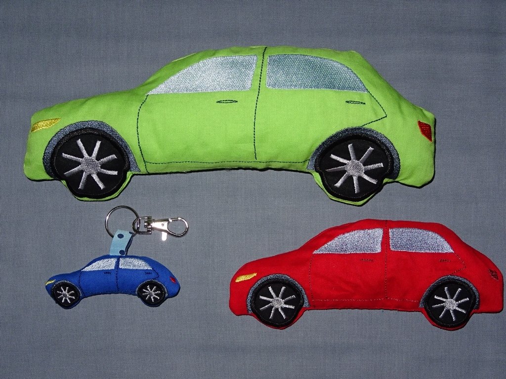 Drei gestickte Auto-Figuren in Grün, Rot und Blau; kleiner blauer Schlüsselanhänger mit Karabiner auf grauem Stoff