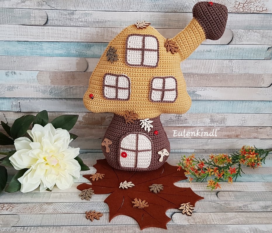 Gehäkeltes Pilzhaus-Kissen in Gelb und Braun mit weißen Fensterapplikationen, Holzknöpfen und Herbstlaub vor Holzpaneelen