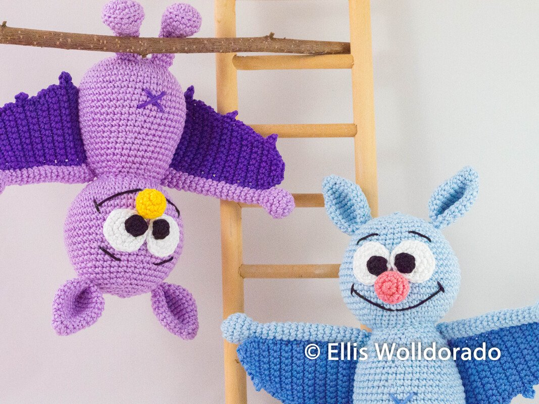 Zwei gehäkelte Amigurumi-Fledermäuse, lila kopfüber hängend und hellblau sitzend vor einer Holzleiter