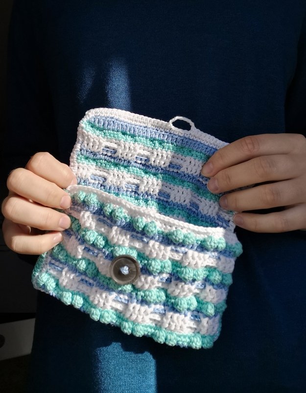 Hände halten ein gehäkeltes Täschchen in Weiß, Blau und Türkis mit Bubbelmaschen und Knopfverschluss