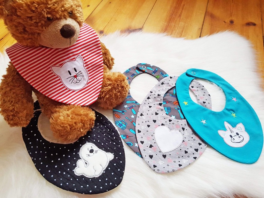 Fünf Baby-Lätzchen mit verschiedenen Applikationen (Katze, Robbe, Einhorn, Herz, Werkzeugmotive) auf hellem Untergrund; ein Teddybär trägt ein gestreiftes Lätzchen.