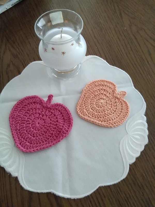 Zwei gehäkelte herzförmige Untersetzer (pink und orange) auf weißem Deckchen neben einer Kerze.
