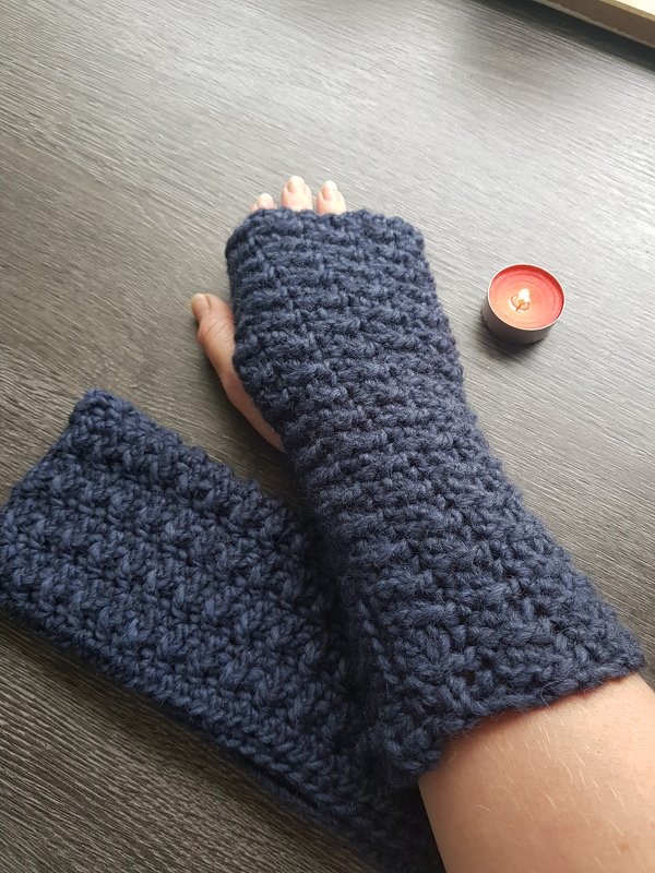 Dunkelblaue gehäkelte fingerlose Stulpe am Unterarm, zweite Stulpe liegt auf dunklem Holztisch neben einem Teelicht