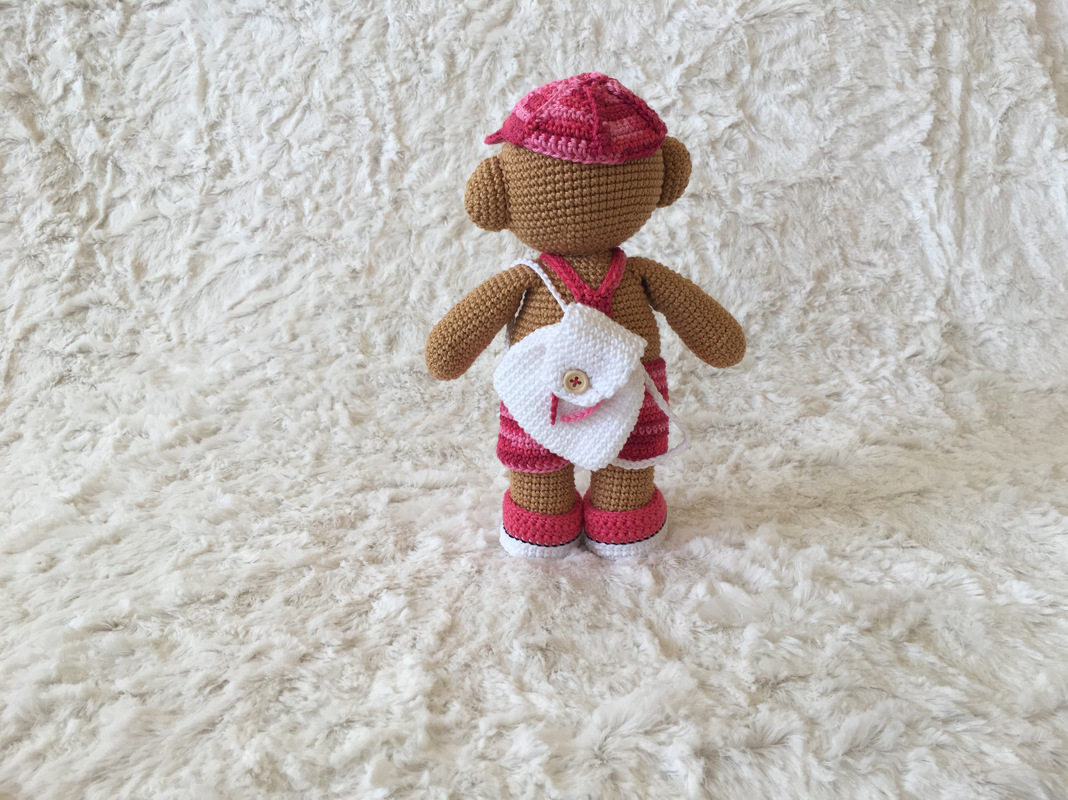 Gehäkelter brauner Teddybär von hinten mit rosa Mütze, weißem Rucksack mit Knopf und rosafarbenen Schuhen auf hellem, flauschigem Untergrund.