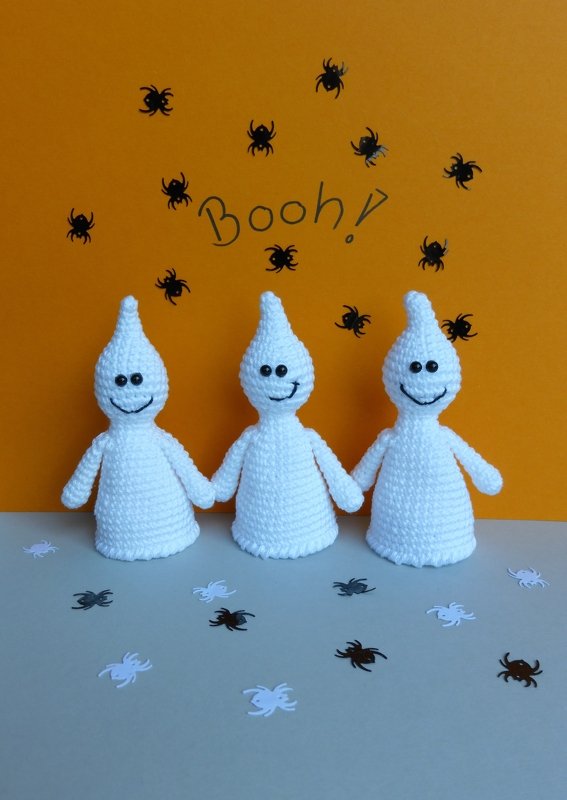 Drei weiße gehäkelte Geister mit lächelnden Gesichtern vor orangefarbenem Hintergrund mit kleinen Spinnen und dem Schriftzug „Booh!“, auf hellgrauem Untergrund mit Spinnenkonfetti.