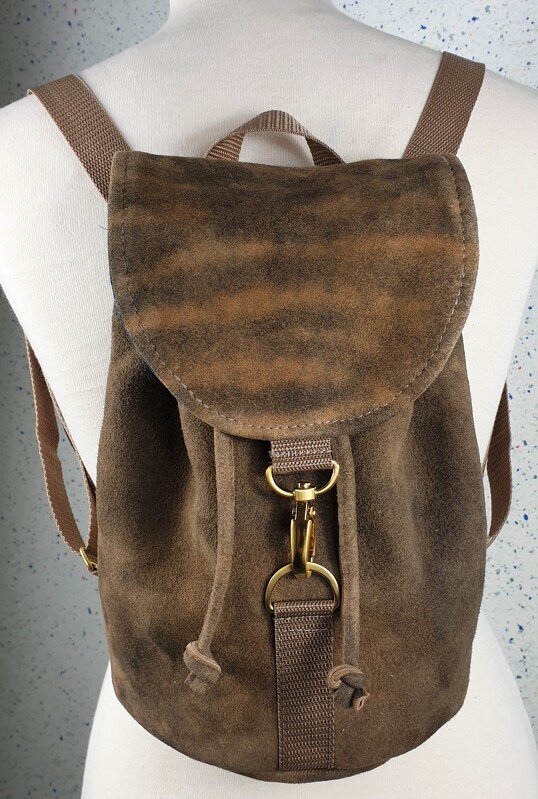 Nahaufnahme eines braunen Rucksacks in genarbter Wildlederoptik mit Kordelzug, Überschlag und messingfarbenen Karabinern auf einer Schneiderpuppe